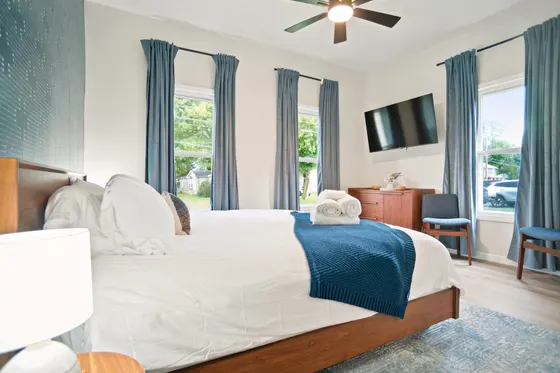 The same king bedroom from the foot of the bed, showing the wall-mounted TV, three windows, and blue-grey curtains.