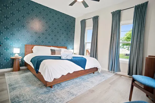 King bedroom with a wood platform bed against a patterned teal accent wall and two curtained windows.