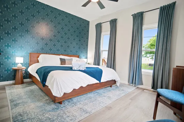 King bedroom with a wood platform bed against a patterned teal accent wall and two curtained windows.