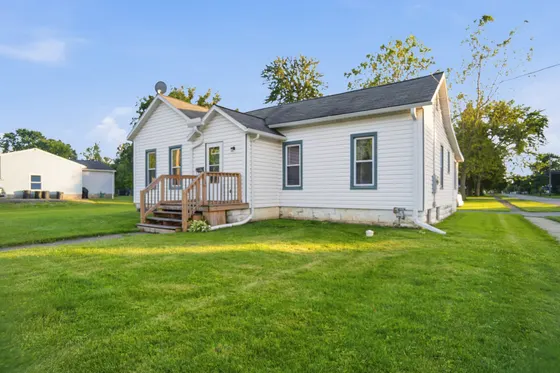 White single-storey cottage with dark green shutters and a small front deck on a green lawn.