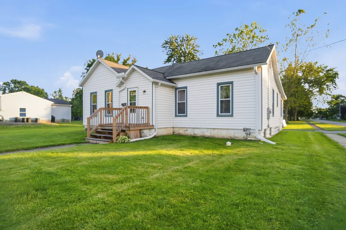 White single-storey cottage with dark green shutters and a small front deck on a green lawn.