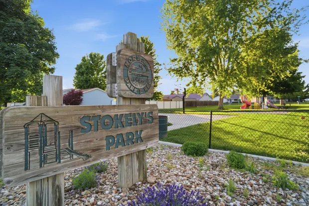 A wooden park sign and playground on a green lawn beneath mature shade trees.