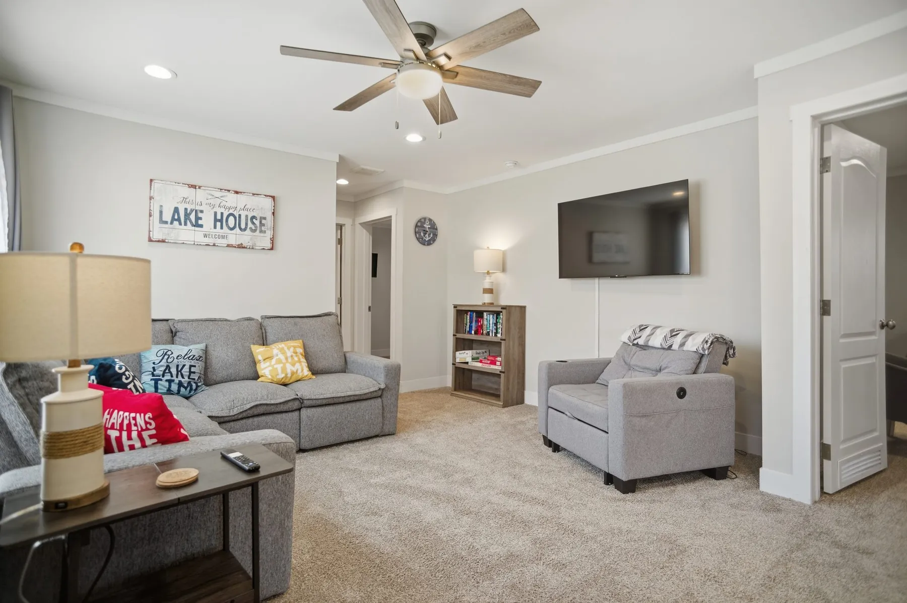 Open living room with a large grey sectional, a wall-mounted TV, and a "Lake House" sign.