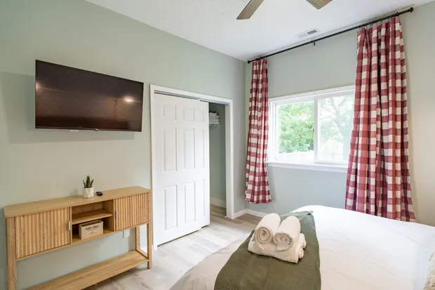 The second bedroom’s wall-mounted TV and dresser beside red gingham curtains.