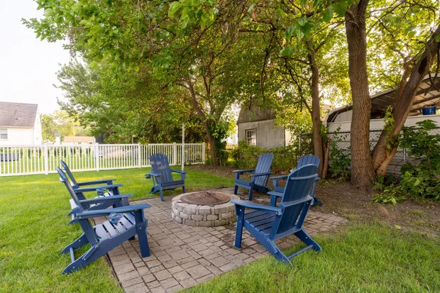 Paver fire pit ringed with blue Adirondack chairs under a shade tree.
