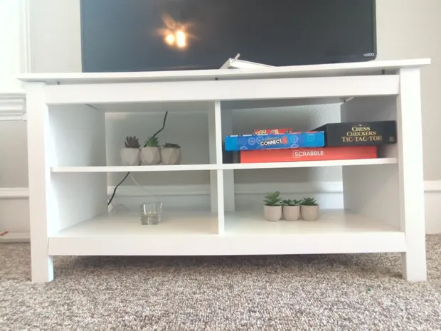 White TV stand with board games and plants on the carpet.